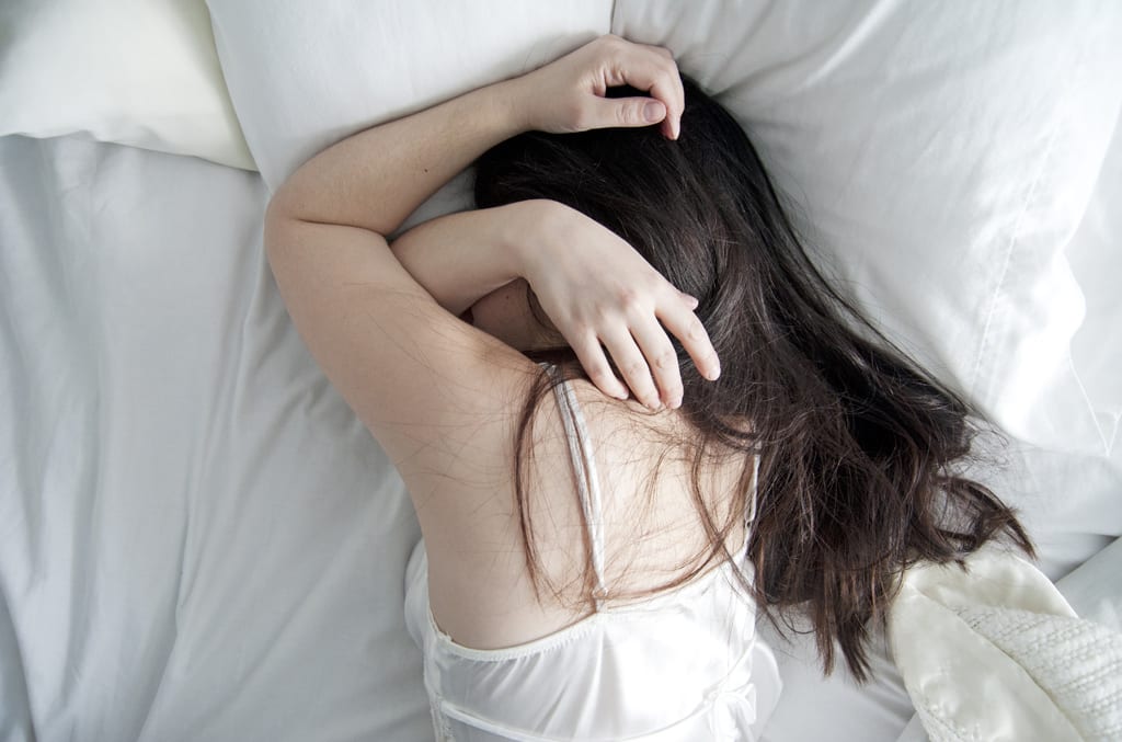 A woman lying on her stomach with her head turned to the side. Her arms are draped over her face in an attempt to block out light.