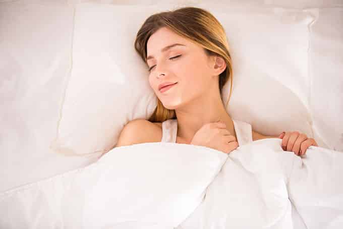 A woman lying on her back, clutching the blanket below her chin and smiling contently