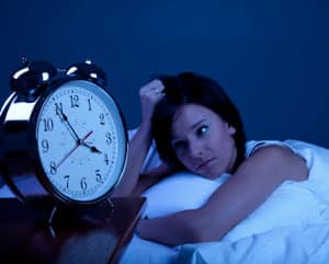 A woman lying awake in a dark room while staring at an alarm clock in the foreground