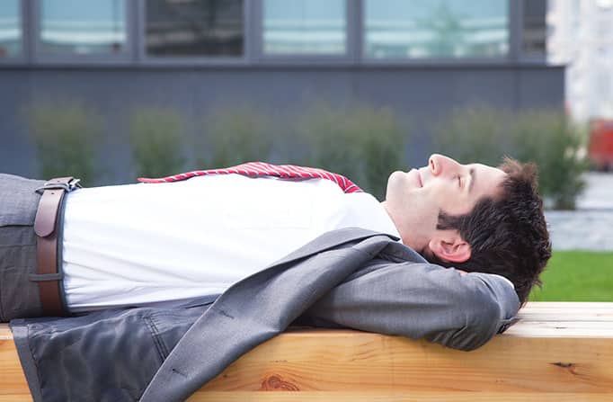 A business man wearing a suit, lying on his back on a wooden bench in an outdoor setting and smiling contently