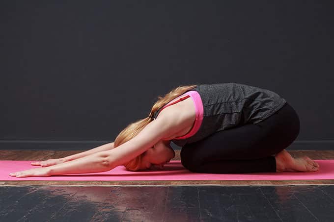 A woman with her legs tucked under her torso as she stretches her hands above her head. Her face is pressed against her yoga mat.