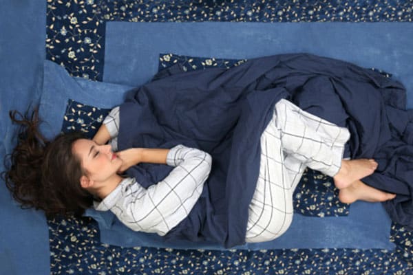 A smiling woman lying in a fetal position on blue bedding, shown from above